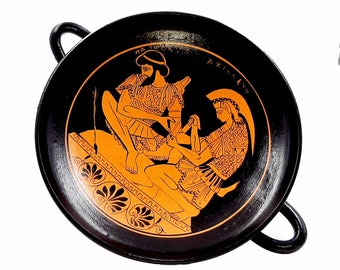 May include: A black and orange Greek pottery plate with two figures in black silhouette. The figures are depicted in a scene of combat, with one figure holding a sword and the other figure holding a shield. The plate has two handles and is decorated with a black and orange border.