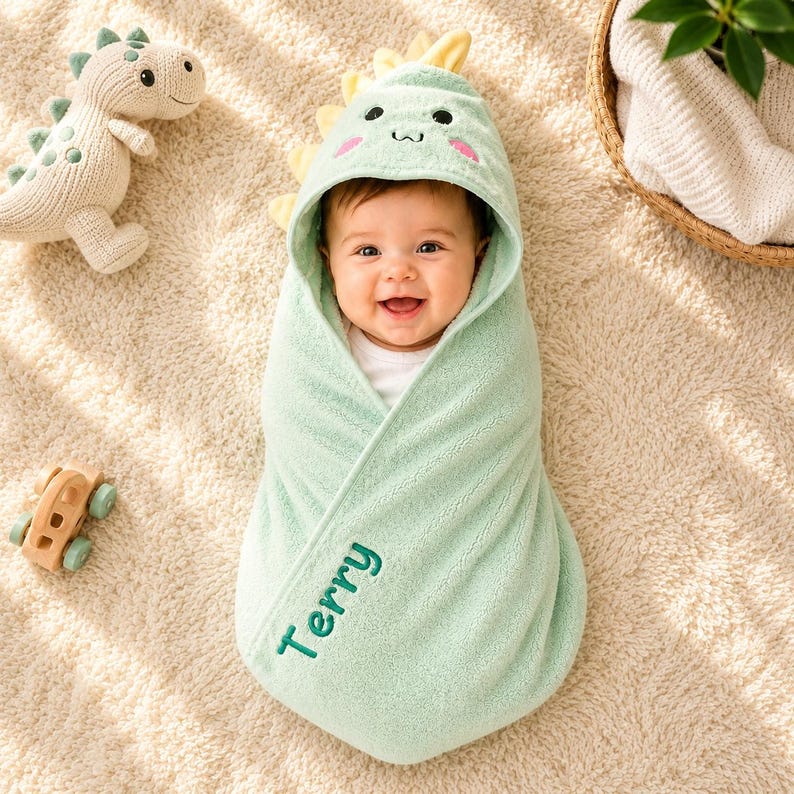 A baby wrapped in a green towel with a dinosaur design, smiling and surrounded by toys on a beige carpet.