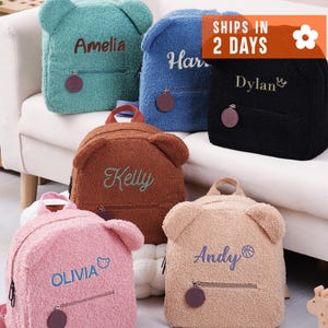 May include: A collection of six fuzzy, bear-shaped backpacks in teal, blue, black, brown, pink, and beige. Each backpack features a front zippered pocket with a circular pull, bear ears on top, and personalized embroidered names and icons.