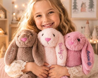 May include: Three plush stuffed bunnies with long floppy ears. The left bunny is brown with the name Harry embroidered on its ear. The center bunny is light pink with the name Becky and a heart. The right bunny is pink with the name Annie and a flower.
