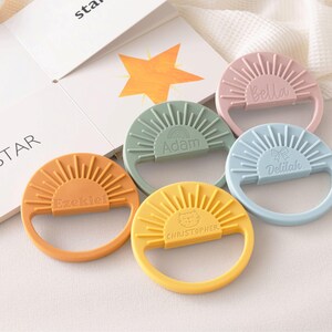 A collection of colorful baby teething rings with names on them, placed on a white surface next to a book.