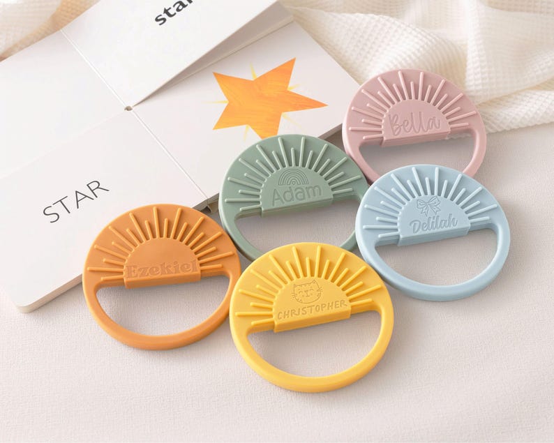 A collection of colorful baby teething rings with names on them, placed on a white surface next to a book.