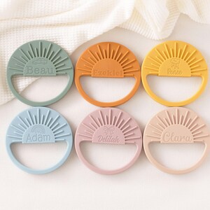A collection of six colorful teething rings, each with a unique name engraved on it, arranged in a grid pattern on a white background.