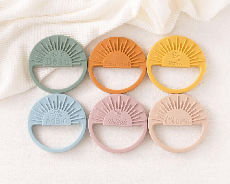 A collection of six colorful teething rings, each with a unique name engraved on it, arranged in a grid pattern on a white background.