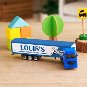 A toy truck is placed on a wooden table, surrounded by various toys and objects, including a toy train, a tree, and a wooden log.