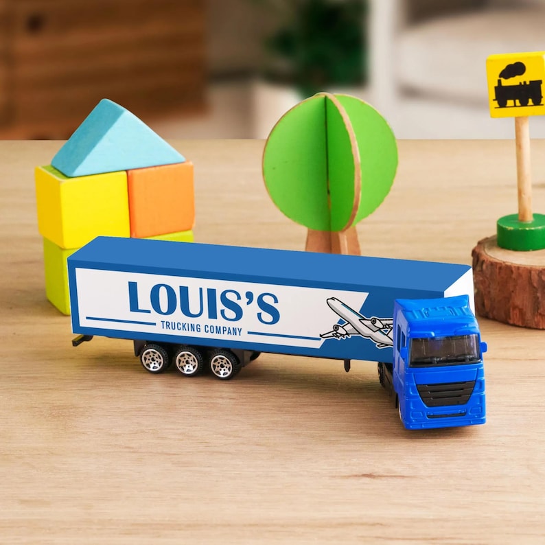 A toy truck is placed on a wooden table, surrounded by various toys and objects, including a toy train, a tree, and a wooden log.