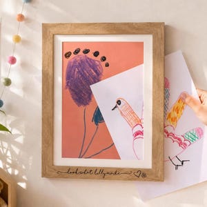 May include: A light wood picture frame with a white mat, featuring the engraved text "look what lilly made" followed by a heart and flower icon. Inside the frame is a child's drawing of a purple flower on a peach background.
