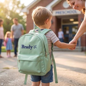 A young boy wearing a green backpack with the name "Brady" on it is being held by an adult, likely a mother, as they walk together on a sidewalk in front of a school building.