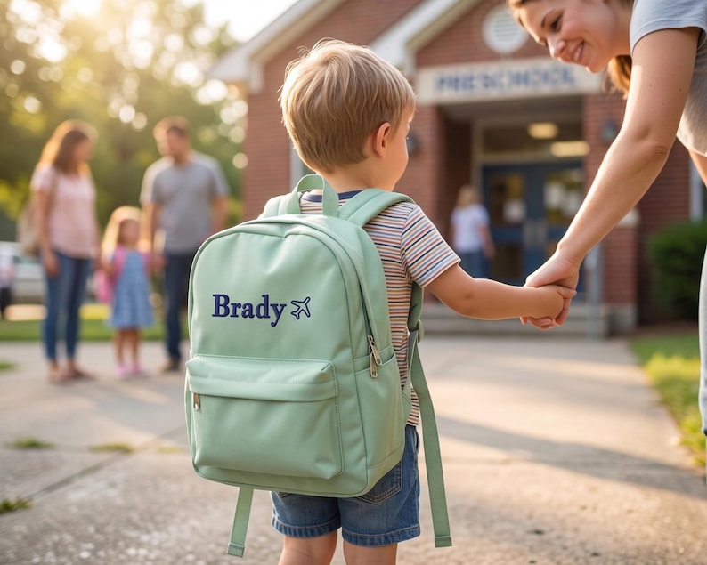A young boy wearing a green backpack with the name "Brady" on it is being held by an adult, likely a mother, as they walk together on a sidewalk in front of a school building.