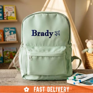 Puede incluir: Una mochila de color verde salvia claro con el nombre Brady impreso en azul oscuro junto a un pequeño icono de avión azul oscuro. La mochila incluye un compartimento principal y un bolsillo frontal con cremallera y tiradores metálicos.