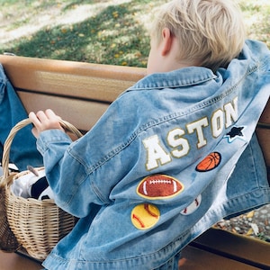 May include: A light blue denim jacket featuring the name ASTON in large white letters with gold trim on the back. The jacket is decorated with embroidered patches of a basketball, a football, a baseball, a softball, and a black star.