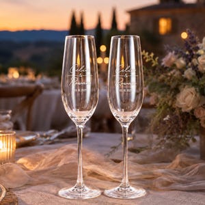 May include: Two clear glass champagne flutes featuring white etched text that reads Celia & Rowan and the date 03.10.2027. The tall, slender glasses have a narrow bowl, a long stem, and a circular base, designed for serving sparkling beverages.