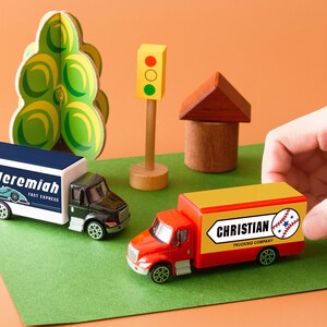 A hand is reaching for a toy truck labeled "Christian" on a green surface, surrounded by other toy vehicles and a miniature toy house.