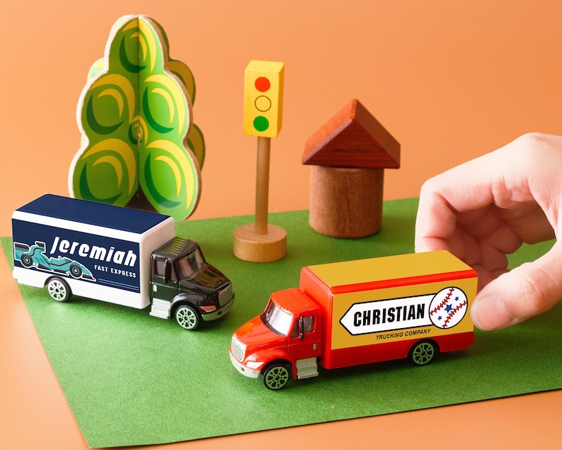 A hand is reaching for a toy truck labeled "Christian" on a green surface, surrounded by other toy vehicles and a miniature toy house.