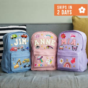 May include: Three backpacks in light blue, pink, and lavender, each featuring embroidered name patches and various colorful fabric appliqués. The blue bag displays 'JIM', the pink bag 'ANNE', and the lavender bag 'MIA' with assorted icons.