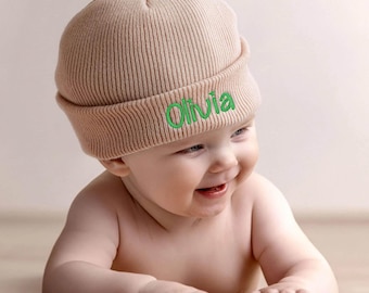 May include: A light beige knit beanie with a folded cuff featuring the name Olivia embroidered in bright green script lettering. The hat is made of a ribbed fabric and is designed to fit snugly, providing a simple and clean look for headwear.