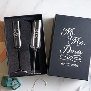 a black box with a white inscription that reads "Mr. & Mrs. Davis" and the date "09. 17. 2026" below it. The box is placed on a marble surface, and there are two champagne flutes inside the box.