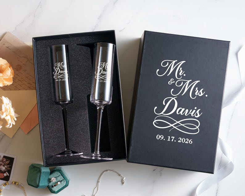 a black box with a white inscription that reads "Mr. & Mrs. Davis" and the date "09. 17. 2026" below it. The box is placed on a marble surface, and there are two champagne flutes inside the box.