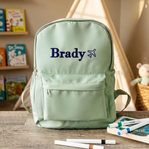 May include: A light sage green backpack featuring the name Brady printed in dark blue text, accompanied by a small dark blue airplane icon. The backpack includes a large main compartment and a smaller front zippered pocket with silver hardware.