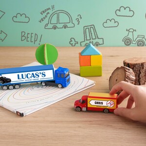 A child's hand is reaching for a toy truck on a wooden surface, with a toy train and other toys nearby. The background features a wall with various drawings and text, including words like "BEEP!" and "VROOM!" and other playful elements.