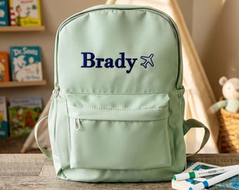 May include: A light sage green backpack featuring the name Brady printed in dark blue lettering, accompanied by a small dark blue airplane icon. The backpack has a large main compartment and a smaller front zippered pocket with a silver zipper pull.