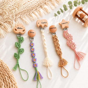 May include: Five macrame pacifier clips with wooden attachments engraved with the names Aiden, Anna, Sophia, Sebastian, and Charlotte. The clips feature various designs including green leaves, purple flowers, cream, tan, and pink knotted patterns.
