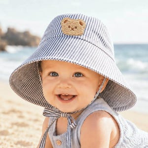 May include: A bucket hat featuring a blue and white vertical striped pattern with a wide brim. The front of the hat is adorned with a small, textured, light brown bear face patch. The hat includes a matching striped chin strap that ties under the chin.