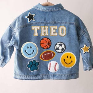 Op de afbeelding: Een lichtblauw spijkerjack met de naam THEO in witte en gouden varsity-letters. De achterkant is versierd met diverse chenille patches, waaronder een blauwe smiley, een gele smiley, een pootafdruk en verschillende sportballen.