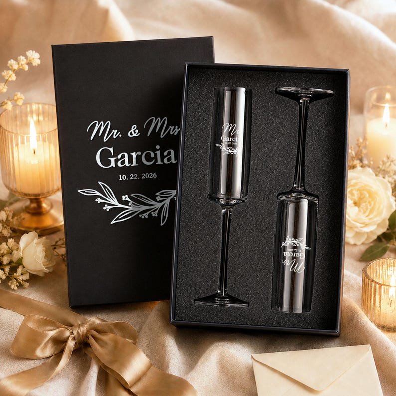 a black box containing two champagne flutes, with a gold ribbon and a white floral design on the box. The box is placed on a table with a white tablecloth, and there are candles and flowers in the background.