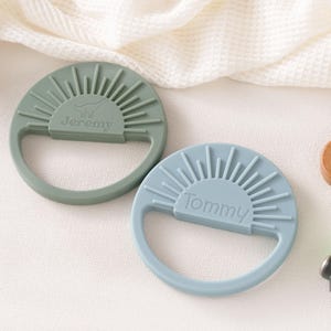 two baby teething toys with personalized names on them, placed on a white surface.