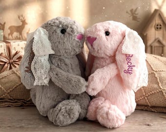 May include: Two plush stuffed rabbits, one grey and one pink, featuring long ears adorned with white lace ribbons. The grey rabbit has the name 'Mona' embroidered on its ear, while the pink rabbit has the name 'Becky' embroidered on its ear.