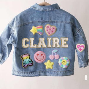 Peut inclure: Une veste en jean bleu clair avec le nom CLAIRE en grandes lettres blanches à bordure dorée. Le dos est orné de patchs brodés colorés, notamment une étoile arc-en-ciel, un cœur rose, un ours en peluche, un visage souriant, des cerises et une fleur.