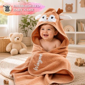 A smiling baby wearing a hooded towel with a deer design, sitting on a rug in a room with a teddy bear and other toys.