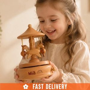 May include: A light brown wooden carousel music box featuring a rotating mechanism with small horse figures suspended from a central canopy. The base is engraved with the name Mia and a small rainbow icon, and it has a small winding knob on the side.