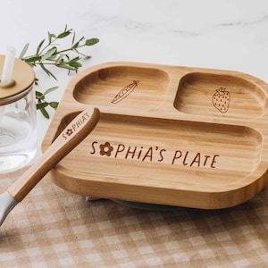 Może przedstawiać: Jasnobrązowy, dzielony talerz bambusowy z trzema sekcjami, wygrawerowany napisem "SOPHIA'S PLATE" i ikoną kwiatka, w zestawie z pasującą bambusową łyżeczką z szarą silikonową końcówką i wygrawerowanym imieniem "SOPHIA'S" na rączce.
