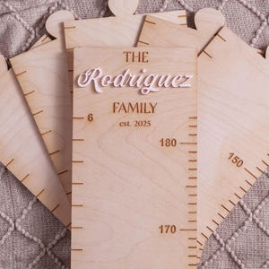 May include: A light-colored wooden growth chart featuring puzzle-piece edges and engraved measurement markings. The center panel displays the text THE Rodriguez FAMILY est. 2025, with the name Rodriguez in a raised white script font.