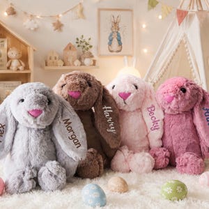 May include: Four plush stuffed bunnies in gray, brown, light pink, and dark pink. Each bunny features long floppy ears with embroidered names: Flex, Morgan, Harry, Becky, and Annie. The bunnies have dark eyes and bright pink noses, sitting upright.