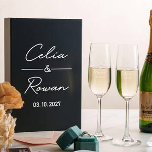 a black box with white text, a bottle of champagne, two champagne glasses, and some flowers on a table.
