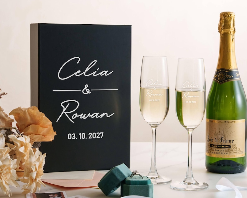 a black box with white text, a bottle of champagne, two champagne glasses, and some flowers on a table.