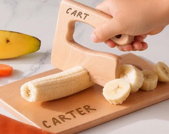 May include: A light-colored wooden toy knife with the word CART engraved on the handle, slicing a peeled banana on a matching wooden cutting board that features the engraved name CARTER on its surface.