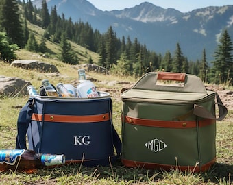 May include: A navy blue cooler bag with the initials KG and an olive green cooler bag with the monogram MGC. Both bags feature brown leather-like trim, top handles, and shoulder straps. Glass bottles are visible inside the open navy blue bag.