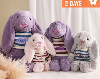May include: Four plush bunny rabbits in shades of purple, pink, and gray. Each bunny wears a striped sweater with a name embroidered on it. The bunnies are sitting on a white surface.