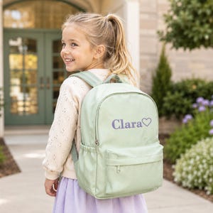 A young girl with blonde hair is smiling and carrying a green backpack with the name "Clara" printed on it.