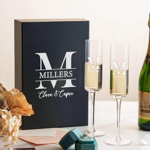 a set of champagne flutes, a bottle of champagne, and a box with a personalized monogrammed logo, all arranged on a table.