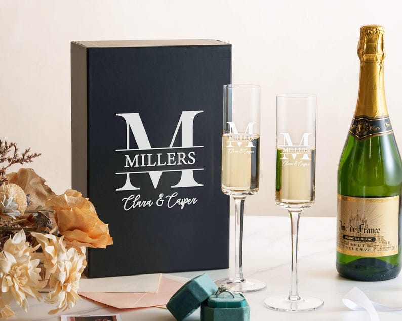 a set of champagne flutes, a bottle of champagne, and a box with a personalized monogrammed logo, all arranged on a table.