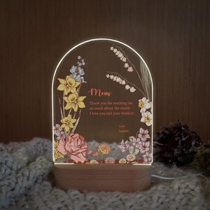 May include: An arched acrylic plaque with a light-up wooden base featuring floral illustrations of yellow daffodils and pink roses. The text reads: Mom, Thank you for teaching me so much about the world. I love you and your wisdom. Love, Isabella.