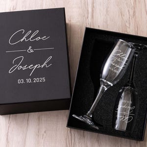 May include: A black gift box with white script reading "Chloe & Joseph" and the date "03.10.2025". The box contains two champagne flutes, also inscribed with the names and date, resting on black foam.