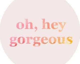 May include: A circular sticker with a pale pink background featuring the text oh, hey gorgeous in a watercolour gradient of pink, orange, and yellow. The lettering is in a lowercase serif font centred on the circular shape.