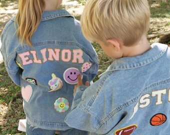 May include: Two light blue denim jackets featuring personalized patches on the back. One jacket displays the name ELINOR in pink letters with various patches including a unicorn, a smiley face, and hearts. The other jacket shows sports patches.