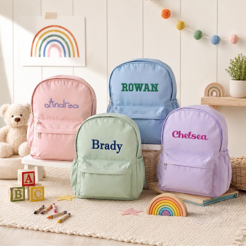 four colorful personalized backpacks, each with a different name written on it, arranged on a rug in a room with a teddy bear and children's toys.